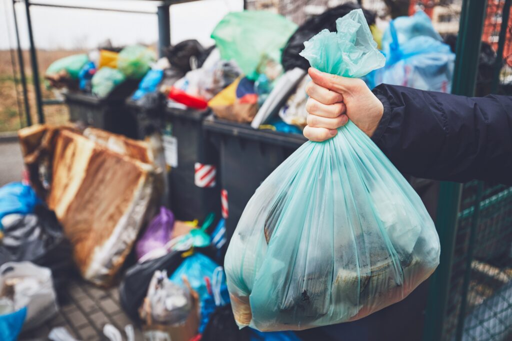 Person hält vollen Müllsack in der Hand und steht neben mehreren gefüllten Mülltonnen