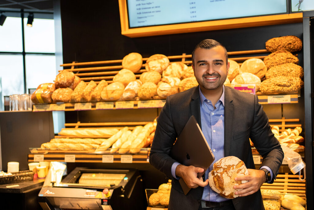 Eyüp Aramaz hält Brot und Laptop in der Hand und steht in einem Bäckereibetrieb