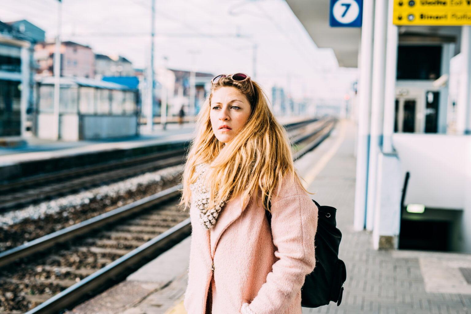 Frau mit einem rosa Mantel und blonden langen Haaren steht an einem Bahnsteig