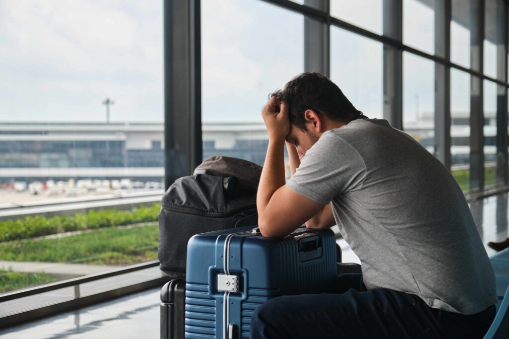 Mann sitzt am Flughafen vor einem Koffer und stützt seine Arme auf seinen Kopf