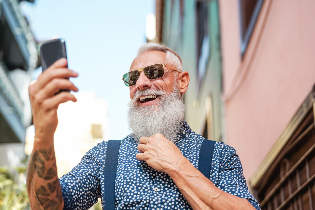 Älterer Mann mit langem weißen Bart und Sonnenbrille trägt ein buntes Hemd und Hosenträger und hält ein Handy lachend mit der rechten Hand nach oben