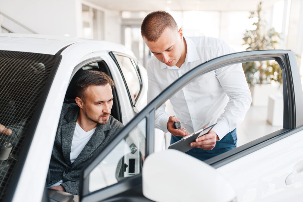 Verkäufer mit weißen Hemd hält eine Unterlage in der Hand sowie einen Autoschlüssel und zeigt diese einem Kunden der in einem weißen Auto sitzt