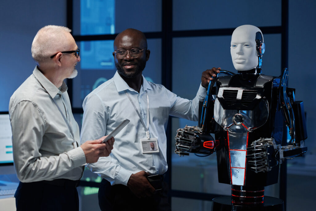 Zwei Männer stehen neben einem Roboter. Ein Mann legt seine Hand auf den Roboter