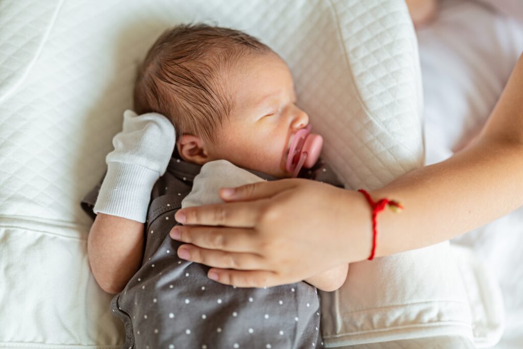 Baby liegt in einem weißem Polster und wird von einer Hand mit einem roten Armband gehalten. Das Baby hat einen rosaroten Schnuller und weiße Handschuhe an
