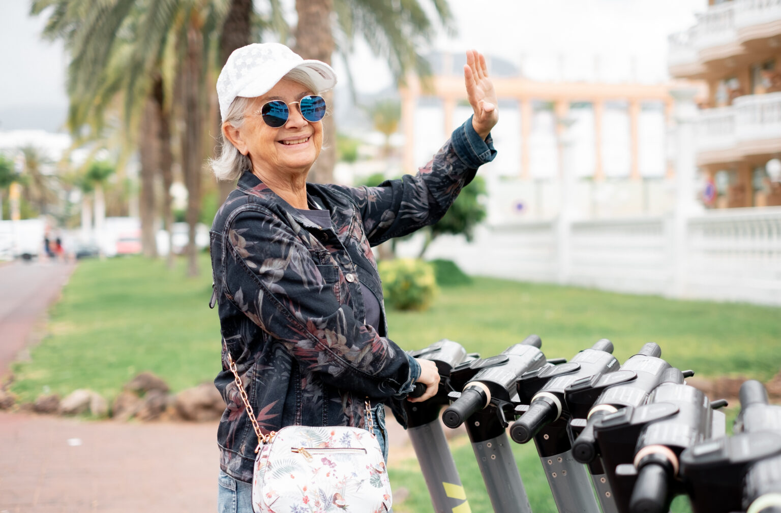 Ältere Dame mit weißer Kappe und Jacke winkt mit einer Hand und steht bei einigen E-Scootern