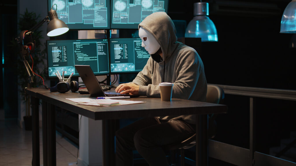 Mensch mit grauem Sweaterpullover vor einem Bildschirm. Der Mensch trägt die Kapuze und eine weiße Maske vor dem Gesicht. Neben ihm viele Monitore