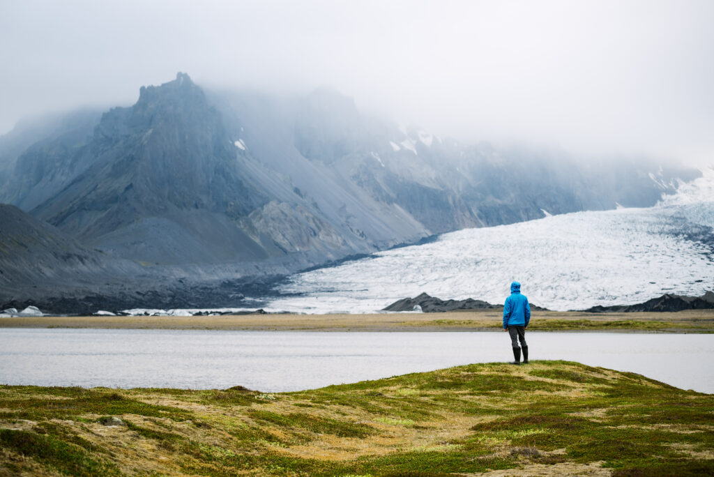 Mensch mit blauem Anorak steht auf einer Wiese und blickt auf einen Gletscher