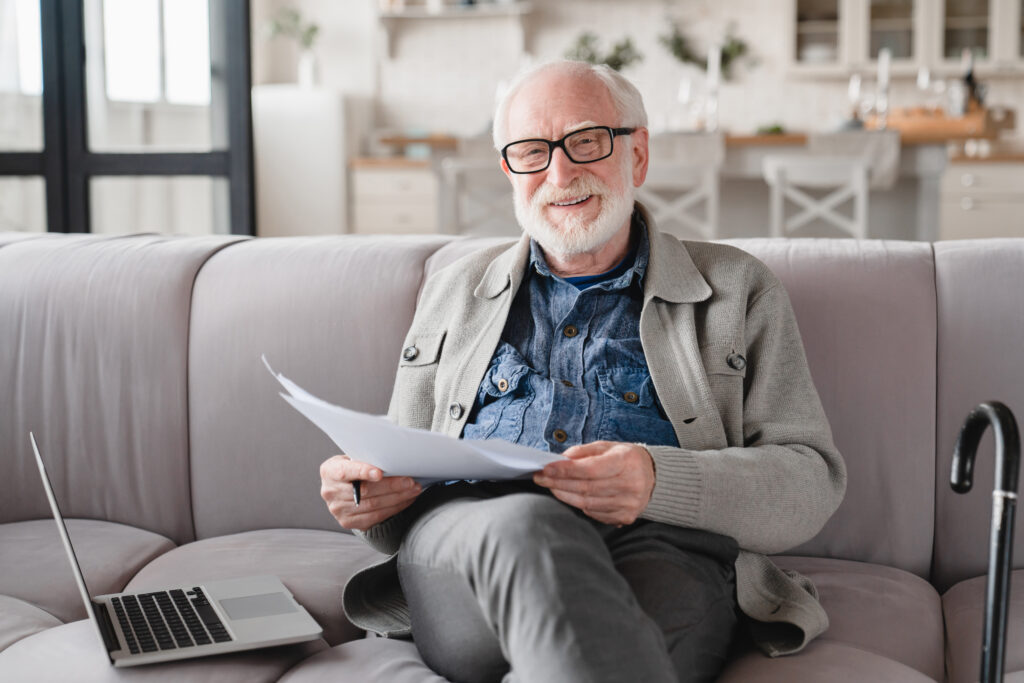 älterer Mann sitzt auf einem Sofa neben einem Computer und hält eine A-4-Zettel in der Hand