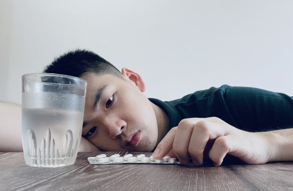 Kind stützt den Kopf auf einen Schreibtisch. Davor ein Glas Wasser und Tabletten